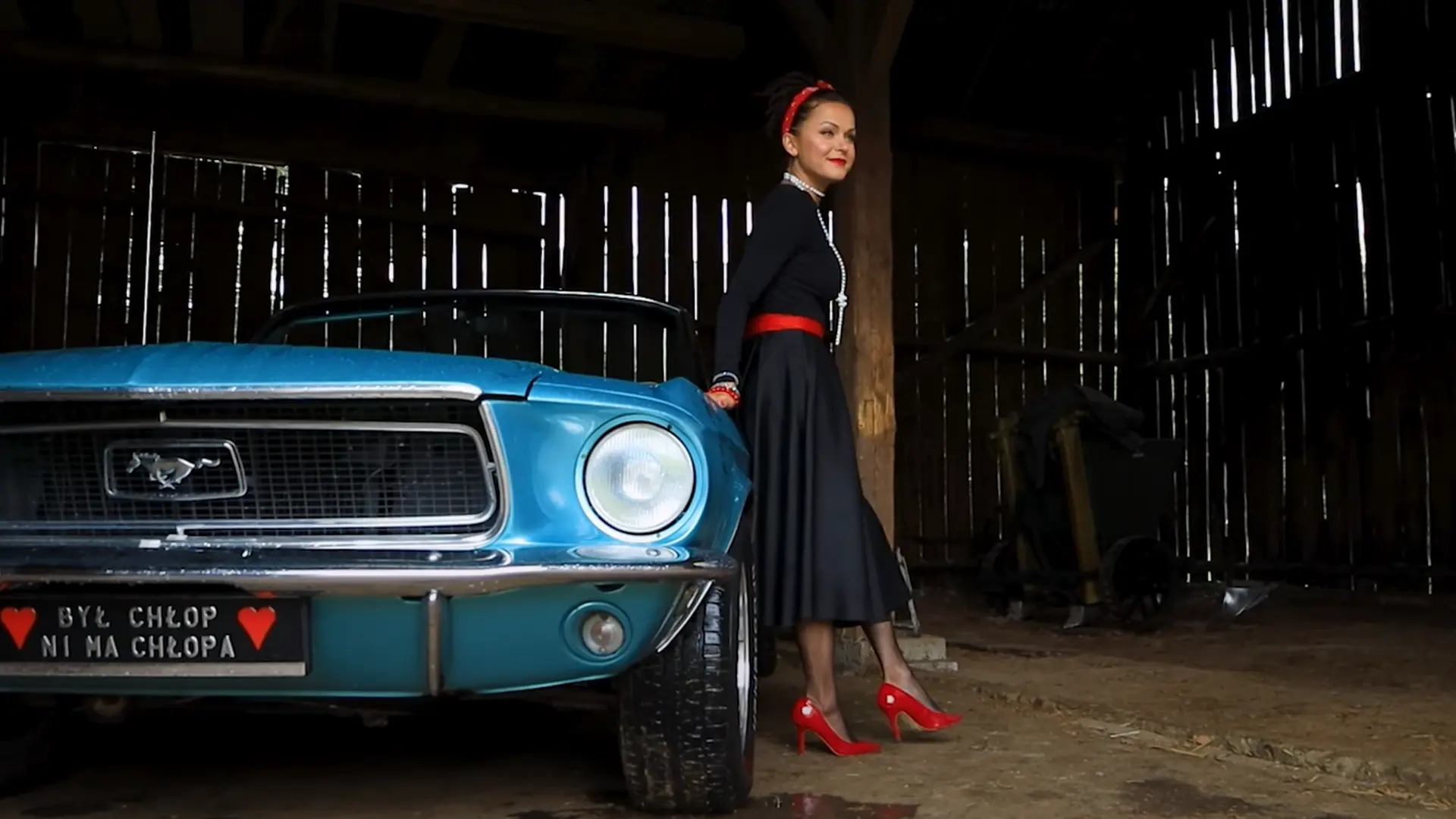 Kadr z backstage'u wideo sesji Pin-Up – stylizowana modelka w czerwonych szpilkach stoi obok błękitnego Forda Mustanga w ciemnej, drewnianej stodole